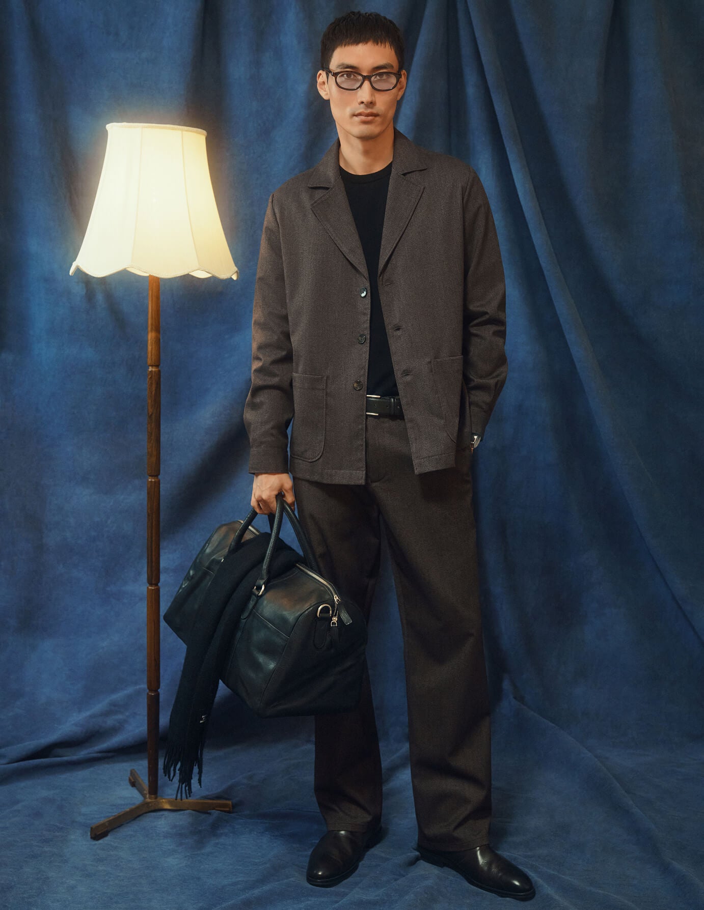 Eine Person, die ein dunkelbeiges Atelier Overshirt und eine dazu passende Hose aus gebrochenem Twill trägt, hält eine schwarze Reisetasche in der Hand.
